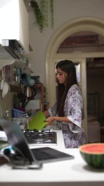 Bir kadın modern bir mutfakta dizüstü bilgisayar kullanıyor ve karpuz kesiyor. Teknoloji ve yemek pişirme arasında bir denge yaratıyor.