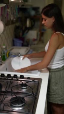 Geceleri modern bir mutfak lavabosunda bulaşık yıkayan bir kadın, bir soba ve çeşitli mutfak malzemeleriyle birlikte..