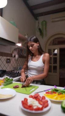 Beyaz atletli genç bir kadın modern bir mutfakta yeşil bir kesme tahtasıyla taze sebze doğruyor..