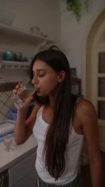Günlük kıyafetli genç bir kadın mutfağında su içiyor, sağlıklı bir yaşam tarzı ve refahı teşvik ediyor.