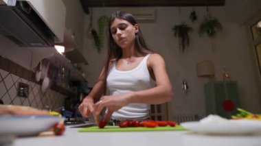 Genç bir kadın, yeşil bir kesme tahtasında sebze doğrayarak modern bir mutfakta yemek hazırlıyor..