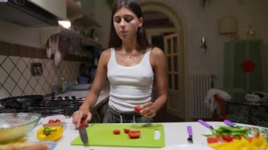 Genç bir kadın, yemek hazırlarken, sıradan bir tişört giyip, modern bir mutfakta sebze doğruyor.