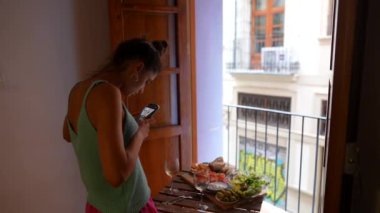 Bir kadın güneşli bir balkonda yemeğini çekerken akıllı telefonunu kullanır, günün ve manzaranın tadını çıkarır.