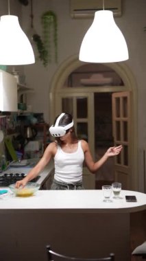 Mutfakta yemek pişirirken, teknoloji harmanlarken ve günlük rutin işler yaparken VR kulaklık takan genç bir kadın..