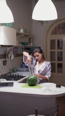 Modern bir mutfakta, bir kadın etrafı çeşitli mutfak malzemeleriyle parlak ışıkların altında karpuz kesmekle meşgul.