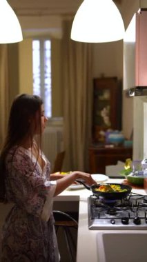 Genç bir kadın modern bir mutfakta yemek pişiriyor. Doğal ışık ve rahat bir atmosferi var. Yemek hazırlıyor.