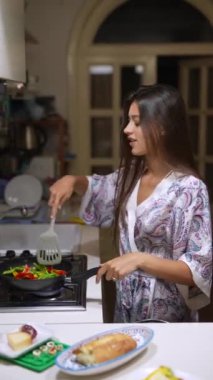 Şablonlu bir bornoz giymiş genç bir kadın, mutfak faaliyetleri sırasında modern bir mutfakta yemek pişirirken görülüyor.