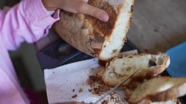Ahşap bir masada ev yapımı ekmek dilimleyen birinin yakın plan görüntüsü kırıntıları ve çıtır çıtır bir kabuğu ortaya çıkarıyor.
