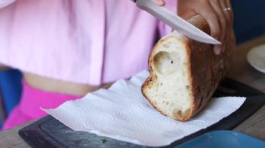 Pembe önlüklü bir kadın ekmek bıçağıyla taze ekmek kesiyor ve kağıt havlunun üzerinde dinleniyor.