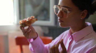 Gözlüklü ve pembe tişörtlü genç bir kadın kapalı bir ortamda bir dilim pizza yiyor.