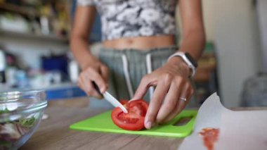 Saati olan bir kadın yeşil bir kesme tahtasında domates dilimliyor, yemek pişirme ve sağlıklı beslenme alışkanlıklarını vurguluyor.