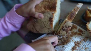 Bir yakın plan, yeni yapılmış ekmeği dilimleyen elleri, etrafa saçılmış kırıntılarla birlikte ahşap bir kesme tahtasıyla yakalıyor.