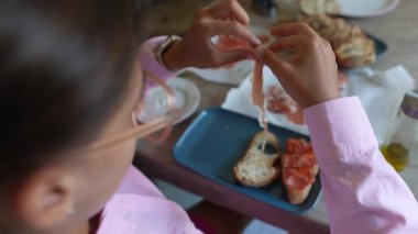 Pembe tişörtlü bir adam ince et dilimleri ve sebzeli bir parça ekmek hazırlar..