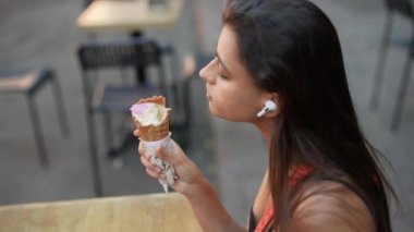 Bir kadın açık hava kafeteryasında oturuyor, AirPod takıyor, modern şehir ortamında dondurma külahının tadını çıkarıyor.