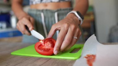 Kırmızı domatesleri yeşil bir kesme tahtasındaki bıçakla kesen ellerin yakın çekimi. Kapalı mutfak ayarları.