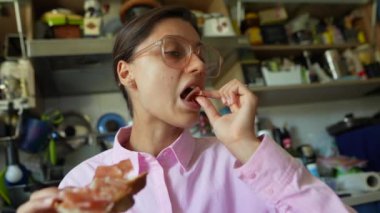 Bardaklı genç bir kadın sıcacık, modern bir mutfakta bir dilim tostun tadını çıkarıyor..