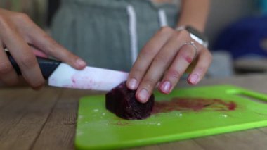 Bir insan yeşil bir kesme tahtasındaki bıçakla pancar kökünü dikkatlice kesiyor. Elleri görünür bir şekilde yakın plan çekimde.