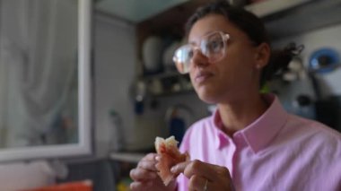 Bulanık mutfak arka planında pembe gömlekli, gözlüklü bir kadın bir şey yerken mutlu görünüyor.