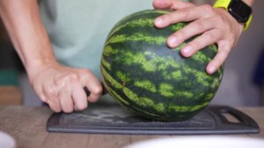 Birisi karpuzu bıçakla kesiyor. Mutfaktaki bir kesme tahtasının üzerinde. Ev ortamında saat takıyor.