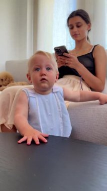 Bir anne ve bebeği evdeler. Bebek yakınlarda dururken anne akıllı telefonunu kullanarak kanepede oturuyor.