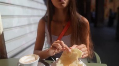 Genç bir kadın açık bir kafede oturmuş güneşli bir günde bir fincan kahveyle bir parça pastanın tadını çıkarıyor.