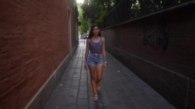 Genç bir kadın dar bir sokakta yürüyor, hayat dolu şehir hayatını ve gençlik enerjisini sergiliyor.