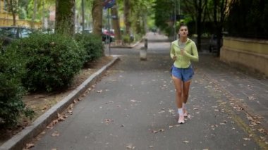 Genç bir kadın, yemyeşil ve park halindeki arabalarla çevrili canlı bir şehir caddesinde yürüyor.