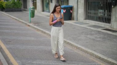 Şık, genç bir kadın işlek caddede güvenli bir şekilde yürüyor, akıllı telefon ekranına derinden odaklanmış.