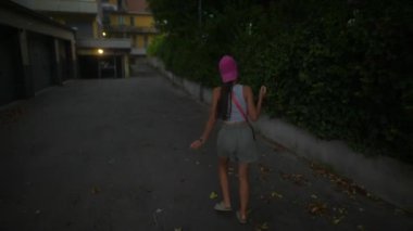 Genç bir kadın alacakaranlıkta, yemyeşil ve yumuşak ışıklarla çevrili bir şekilde şehir dar sokaklarında geziniyor.