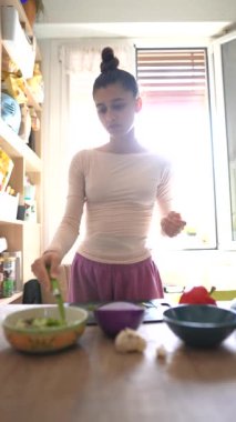 Bir kız, rahat ve parlak bir mutfakta yemek pişirme becerisini gösteren taze malzemeleri ustalıkla hazırlar.