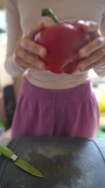 Bir insan canlı, parlak bir mutfakta taze malzemeler hazırlarken elinde parlak kırmızı bir dolma biber tutuyor.