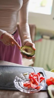 Bir kadın, parlak, havadar mutfağında taze sebzelerle çevrili bir avokadoyu ustalıkla dilimliyor.