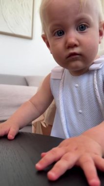 Beyaz elbiseli sevimli bebek, ilginç bir şekilde modern bir oturma odasında siyah masayı keşfediyor.