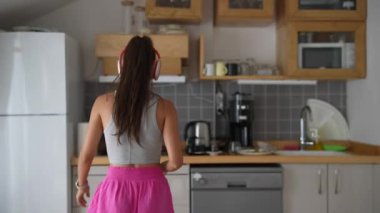 Şık kulaklık takan genç bir kadın, modaya uygun bir mutfakta neşeyle dans ediyor. Enerjiyi ve rahatlığı somutlaştırıyor.