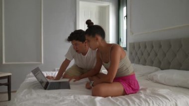 Bir çift genç yetişkin şık bir yatak odasındaki yatağın üzerinde dizüstü bilgisayar kullanarak odaklanmış bir tartışmaya giriştiler..