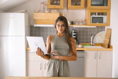 Modern, şık genç bir kadın, çağdaş bir mutfak ortamında otururken aktif olarak laptopuyla meşgul oluyor.