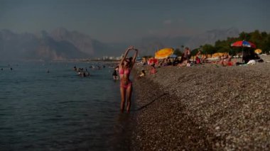 Güneşlenenlerin, yüzücülerin ve arka planda güzel görünen manzaralı dağların olduğu canlı bir sahil sahnesi.