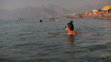 Huzurlu ve sakin bir sahil sahnesi insanların neşeyle yüzdüğü, nefes kesen dağların arka planda olduğu