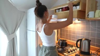 Bir kadın şık bir mutfaktaki raflardan tabakları kapmak için uzanır, ki bu da güzel bir şekilde ev düzenini gösterir.