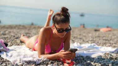 Bir kadın mutlu bir şekilde kumsalda güneşli ve aydınlık bir günün tadını çıkarırken akıllı telefonunu havlunun üzerinde kullanıyor.