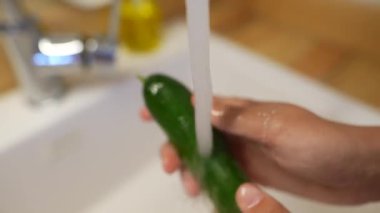 Mutfak lavabosunda, bir insan akan suyun altında taze yeşil bir salatalığı iyice yıkıyor.