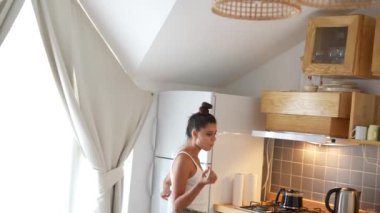 Bir kadın, doğal ışıkla dolu aydınlık ve modern mutfağını keşfederken mutlu bir şekilde atıştırmaktan zevk alır.