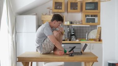 Genç bir adam, modern bir mutfak masasında oturmuş, laptopuna odaklanmış, buharlı bir fincan kahvenin keyfini çıkarıyordu.