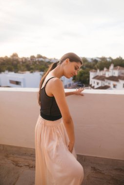 Güzel bir balkonda zarif bir etek giyen zarif bir kadın huzurlu bir günbatımı sahnesinde poz verir.