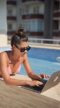 Şık güneş gözlüğü takan hoş bir kadın güneşli bir günde havuz kenarında dizüstü bilgisayarını kullanıyor.