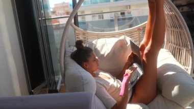 Bir insan rahat bir okuma köşesinde rahat bir şekilde otururken parlak, güneşli bir ortamda dizüstü bilgisayar kullanıyor.