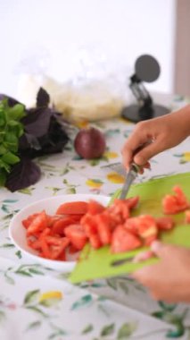 Bir insan maharetle olgun, sulu domatesleri rengarenk bir mutfak masasında kesiyor. Bahçeden taze otlarla süsleniyor.
