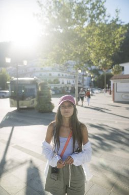 Parlak güneş ışığıyla aydınlatılmış canlı bir şehir ortamında, şık genç bir kadın dışarıda neşeli bir şekilde poz veriyor.