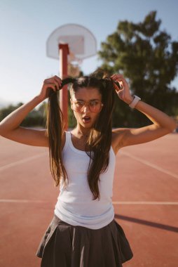 Neşeli bir kız basketbol sahasında güneşli bir günün tadını çıkarıyor, neşeli saç stillerini sergiliyor.