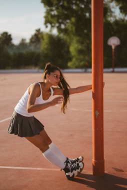Dinamik ve enerjik genç bir kadın spor kıyafetleri içinde bir basketbol direğinin yanında neşeyle kayıyor.
